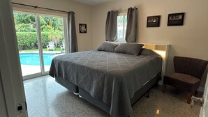 Una camera da letto con un letto, una sedia, un ventilatore a soffitto e vista su una piscina attraverso una porta scorrevole.
