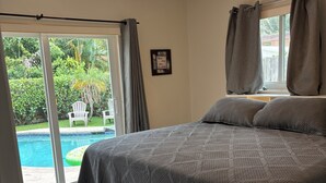 Una camera da letto con un letto, una ventola a soffitto, una finestra con tende e vista su una piscina e sulla vegetazione circostante.