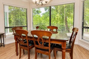 A dining area with a wooden table and chairs, large windows with blinds, and a chandelier.