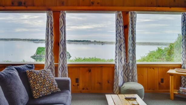 A room with a large window overlooking a body of water, a wooden ceiling, a sofa with patterned cushions, and a small table with a remote control. - Rustic Coastal Smith River Cottage w/ Ocean Views! (Smith River)