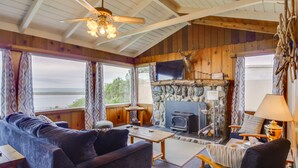 A cozy cabin interior with a stone fireplace, wooden furniture, and a large window overlooking a forested area. - Rustic Coastal Smith River Cottage w/ Ocean Views! (Smith River)