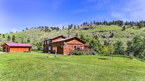 A wooden cabin with a red roof and a large wooden structure nearby, set against a hillside with trees and a clear sky. - South Dakota Vacation Rental Near Mount Rushmore (Hermosa)