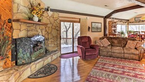 A cozy living room with a stone fireplace, a plush red armchair, a brown sofa, and a large window overlooking a deck. - South Dakota Vacation Rental Near Mount Rushmore (Hermosa)