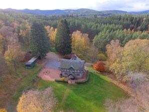An aerial view of a house surrounded by a forest with a well-maintained lawn and a gravel driveway.