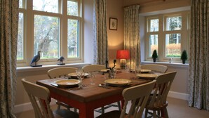 Een eetkamer met een houten tafel gedekt voor zes personen, omringd door witte stoelen. Er zijn twee ramen met gordijnen, een lamp op de tafel en een vogelbeeldje op de vensterbank.