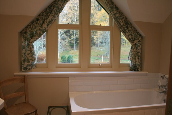A bathroom with a bathtub, a window with floral curtains, and a view of trees outside.