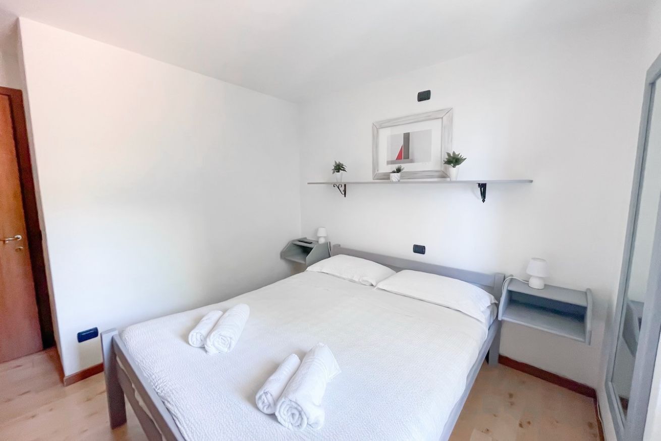 A neatly made bed with white linens and rolled towels, a bedside table with a lamp, a small shelf with decorative items, and a door to the left.
