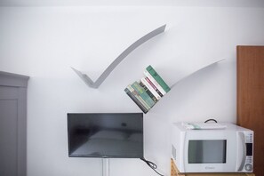 Een flatscreen televisie aan een muur bevestigd, een stapel boeken tegen de muur geleund, en een magnetron op een houten standaard.