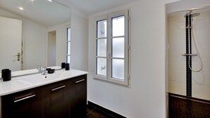 A modern bathroom with a white sink, dark cabinets, a large mirror, frosted window, and a shower with a black tile surround.