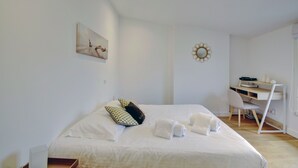 A neatly made bed with pillows and towels, a desk with a chair, a wall-mounted mirror, and a painting.
