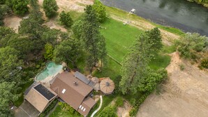 An aerial view of a residential area with a river, a swimming pool, and a house with a large roof and solar panels.