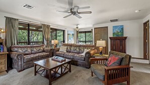 A spacious living room with a large sectional sofa, a wooden coffee table, and a fireplace. There are large windows with curtains, a ceiling fan, and a painting on the wall.