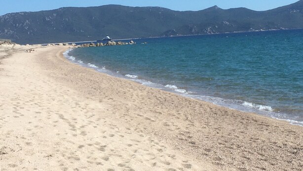 Een zandstrand met kalme golven, een steiger die het water in reikt, en bergen op de achtergrond.