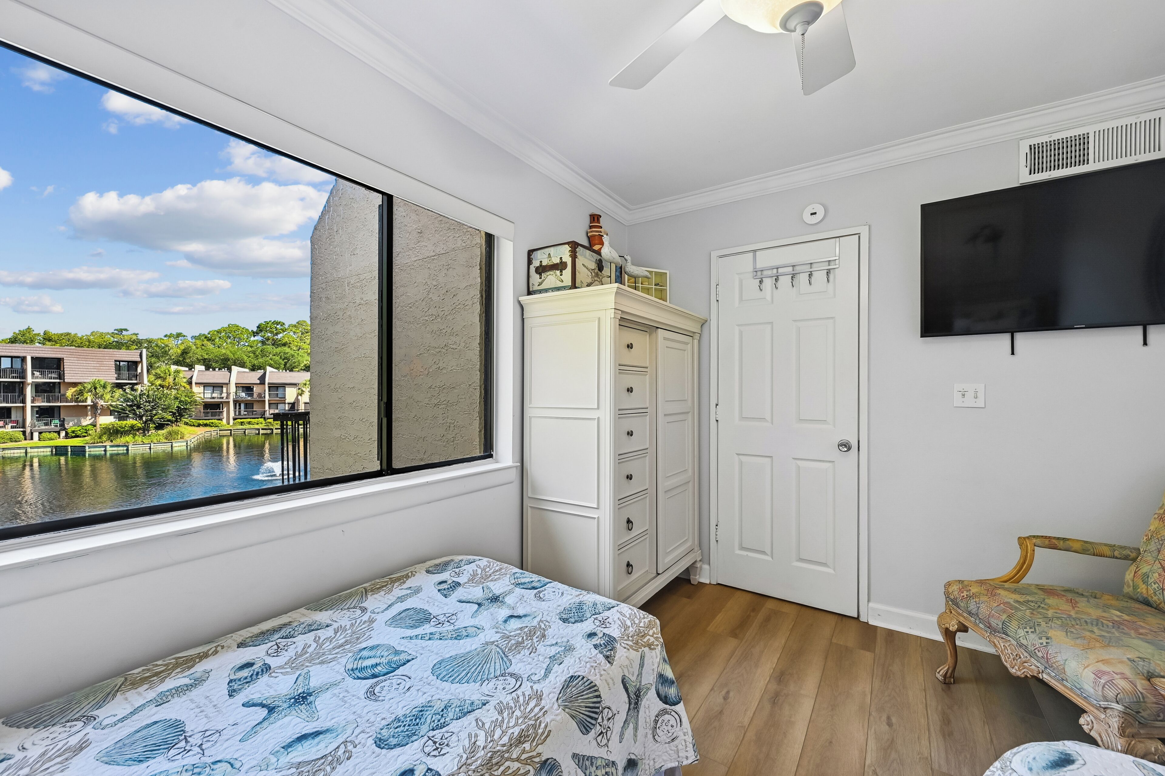 Un dormitorio con un ventanal grande, una cama con cubrecama de temática náutica, un armario blanco, una silla de madera y un televisor de pantalla plana en la pared.