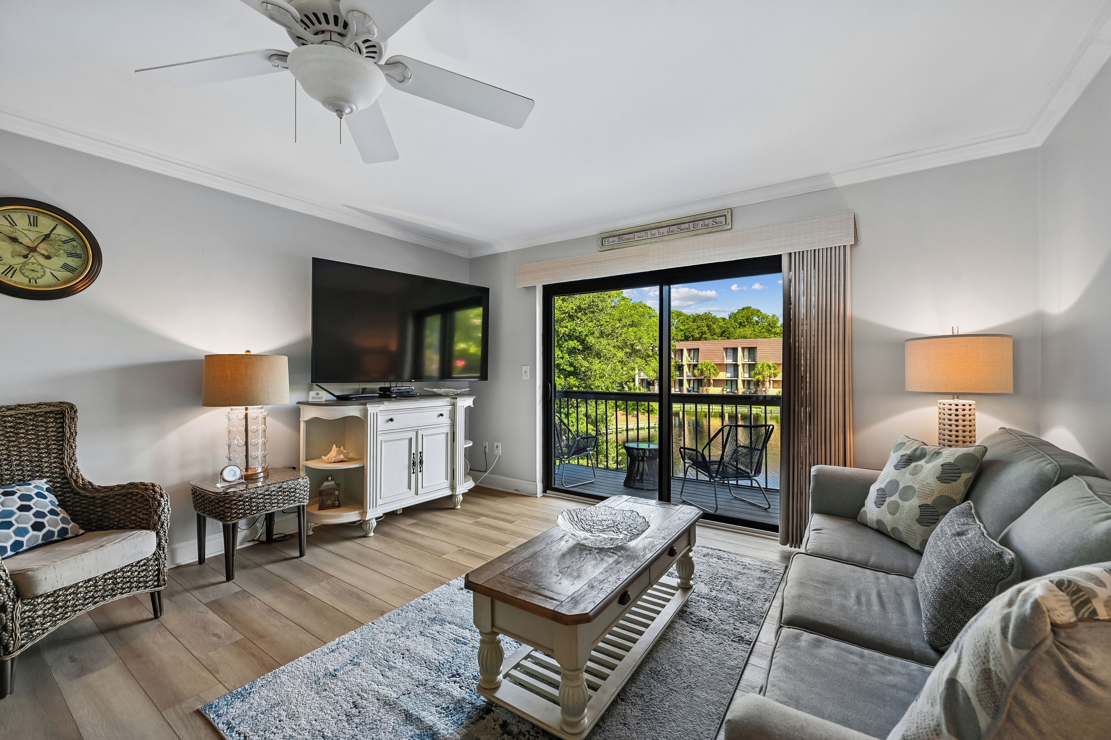 Ein Wohnzimmer mit Deckenventilator, einem Flachbildfernseher, einer Couch, einem Couchtisch und Blick auf einen Balkon mit Gartenmöbeln.