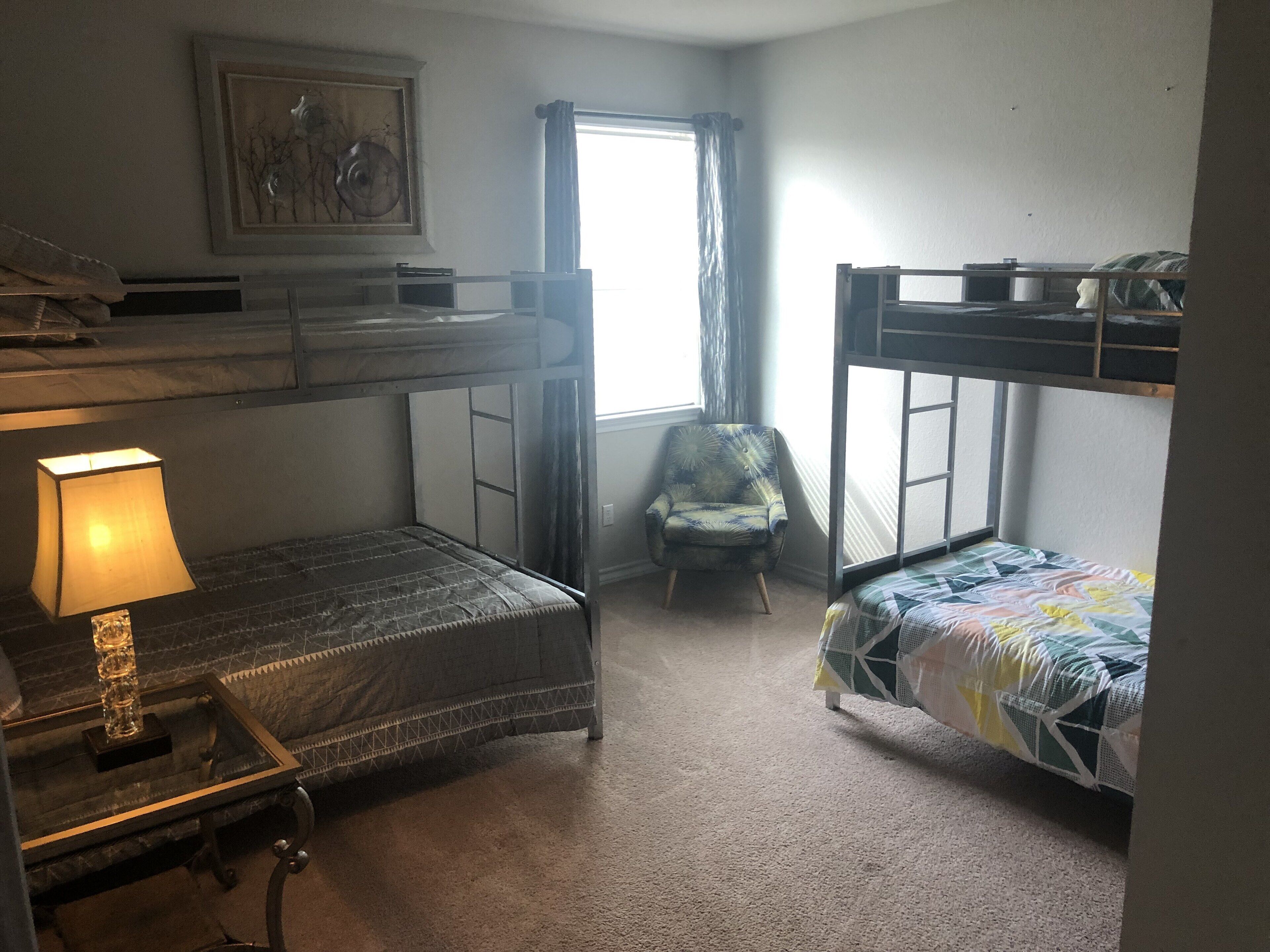 A dormitory room with bunk beds, a desk with a lamp, a chair, and a window with curtains.