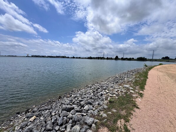 Vista de un lago con orilla rocosa, un sendero arenoso y edificios a la distancia.
