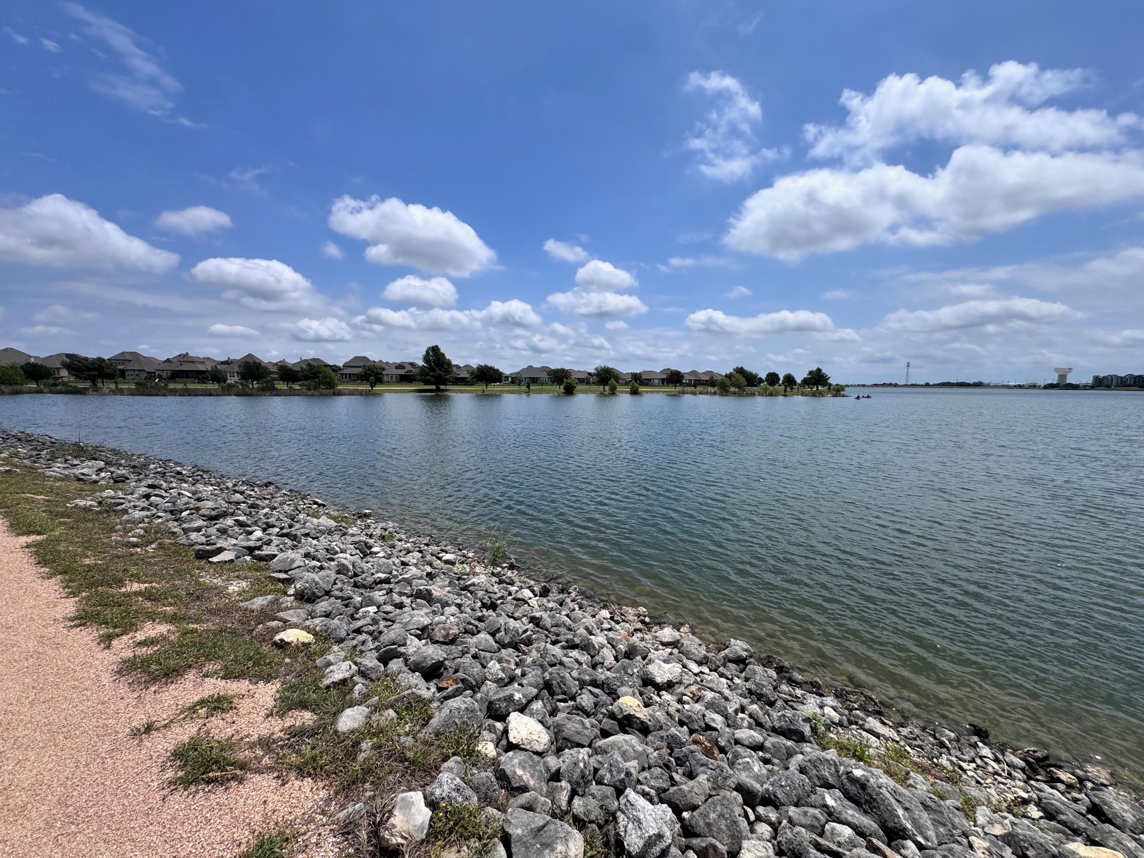 Vista de un lago con un camino de grava, agua tranquila y casas a lo lejos.