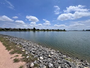 Vista de un lago con un camino de grava, agua tranquila y casas a lo lejos.