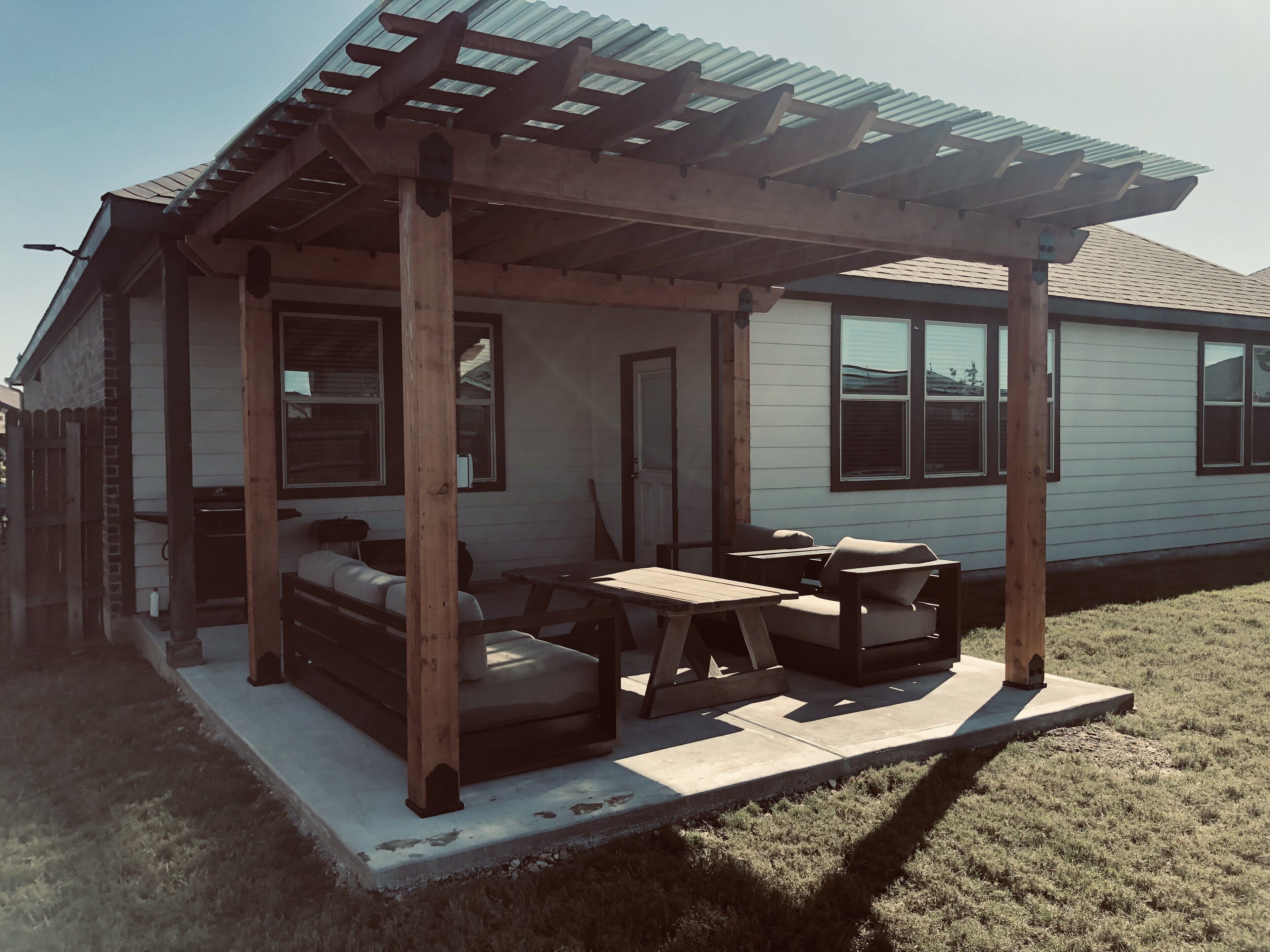 Un patio techado con mobiliario de madera, piso de concreto y una casa al fondo.