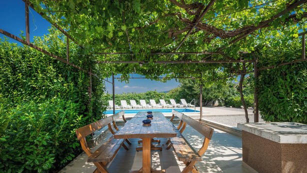 An outdoor dining area with a long wooden table and benches, surrounded by lush greenery and a view of a swimming pool.