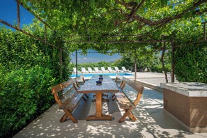 An outdoor dining area with a long wooden table and benches, surrounded by lush greenery and a view of a swimming pool. - Modern villa XXL Makarska (Katuni)