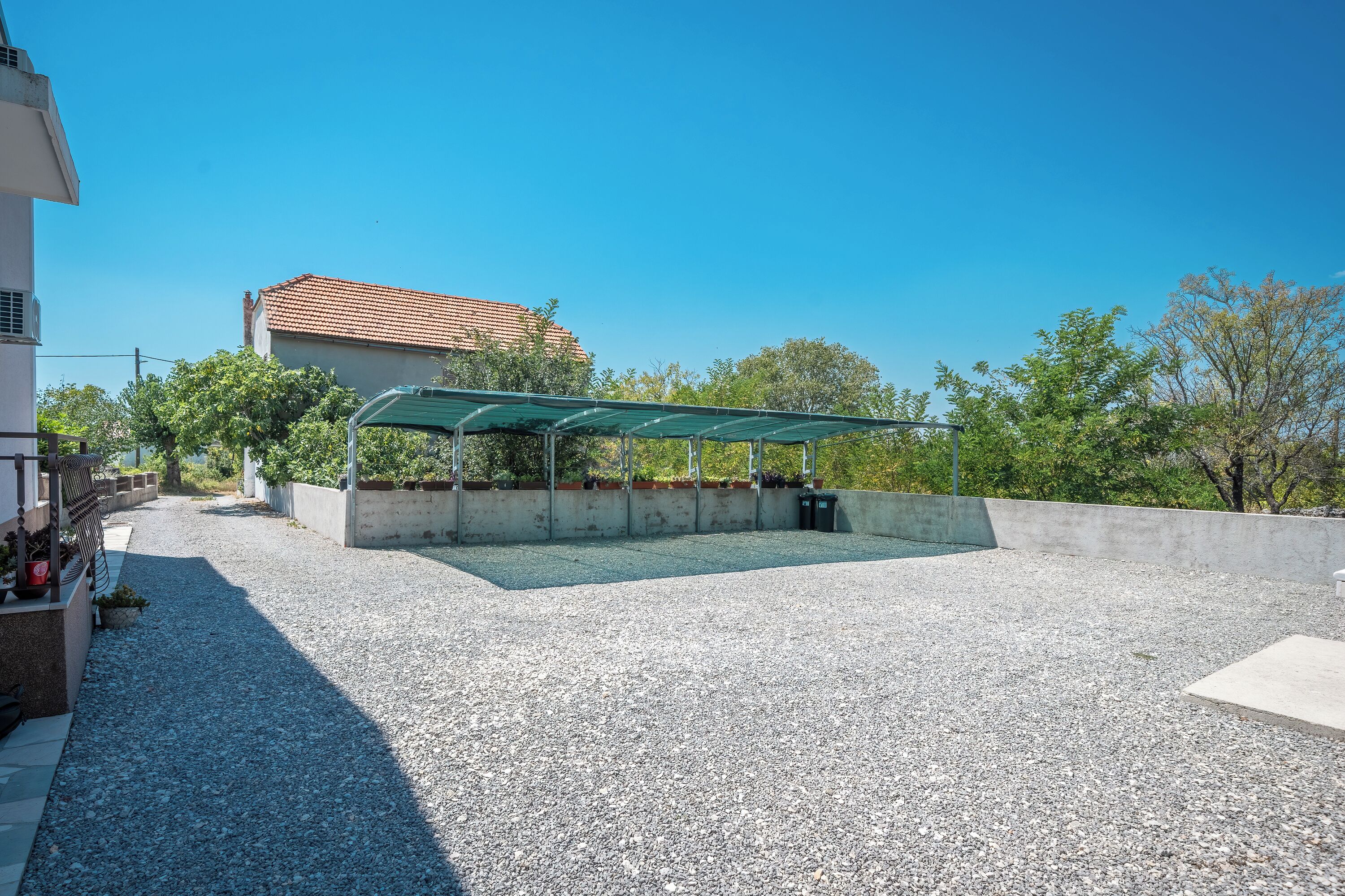 A covered outdoor seating area with a gravel ground, a concrete wall, and a building with a tiled roof in the background.