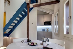 Un comedor con una mesa dispuesta para dos, una escalera azul y una cocina al fondo.