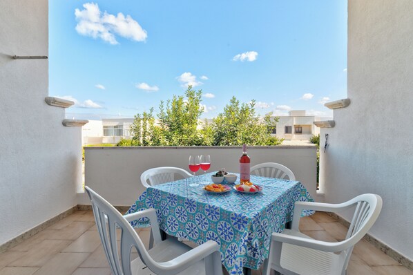 A balcony with a table set for two, white plastic chairs, a tablecloth with a blue and white pattern, and a bottle of red wine.