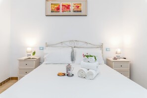 A neatly made bed with white linens, two bedside tables with lamps, a framed picture above the bed, and a small table with toiletries.
