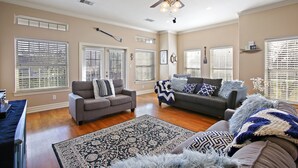 A spacious living room with hardwood floors, two sofas, a ceiling fan, and large windows with blinds. - Boardwalk Home One Block to Kemah Activities  (kemah)
