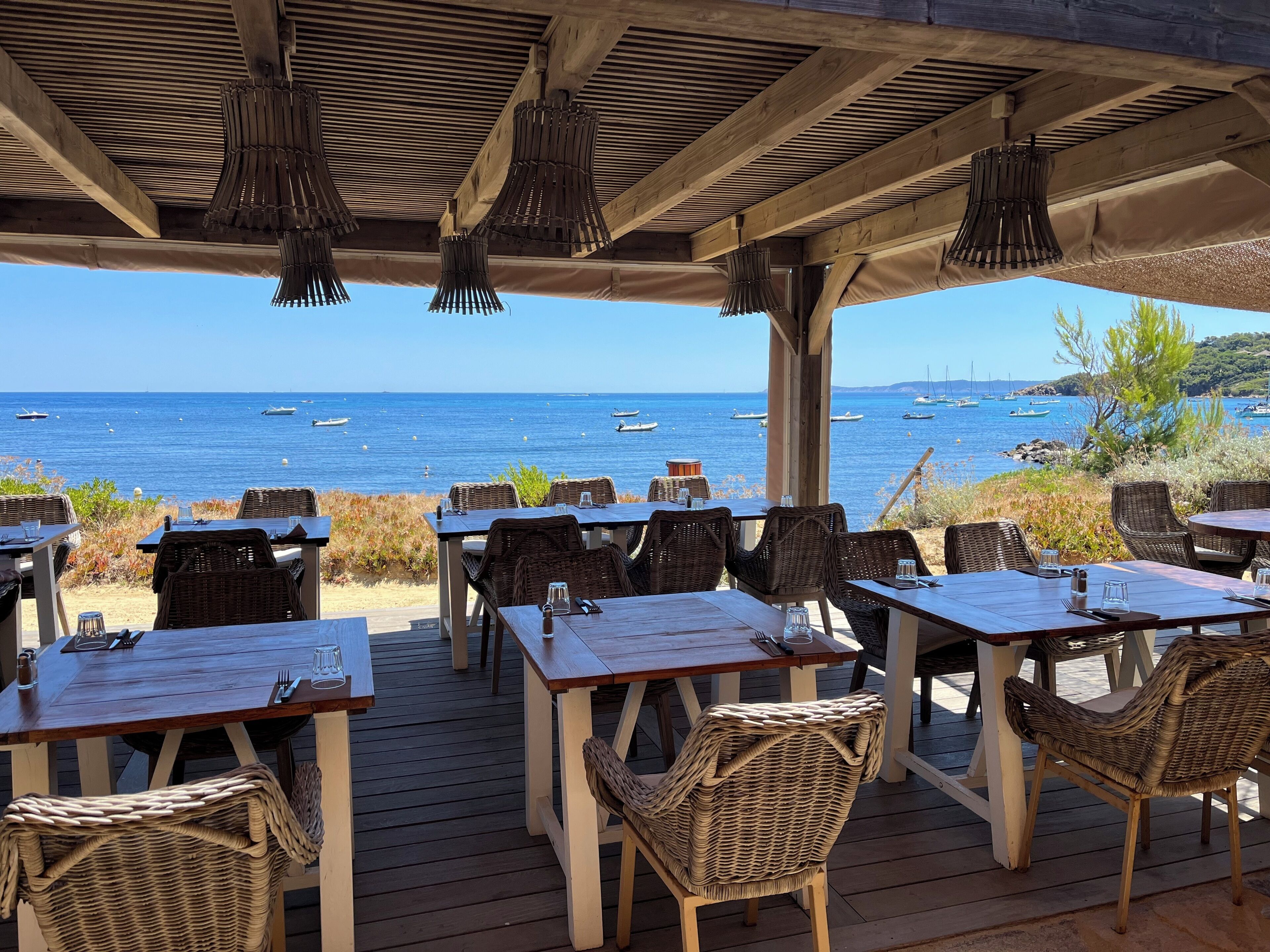 Un espace de restauration en plein air, avec des tables et des chaises en osier, donnant sur une plage où sont amarrés des bateaux.