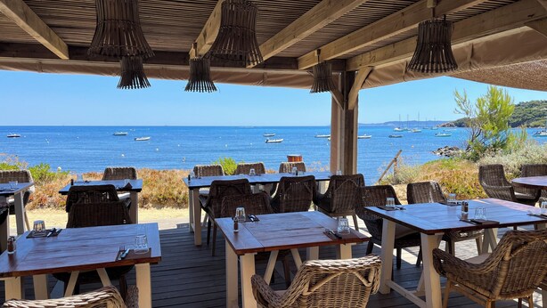 Un área de comedor al aire libre con mesas y sillas de mimbre, con vista a una playa y barcos.