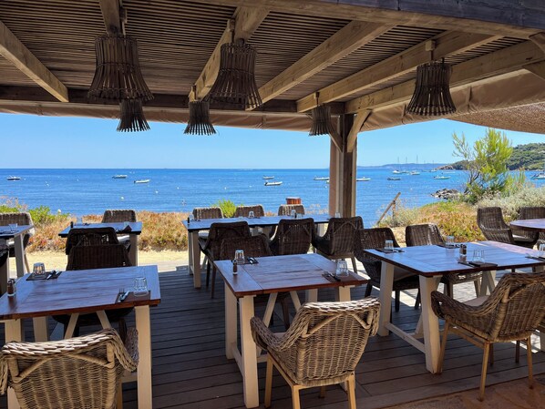 An outdoor dining area with tables and wicker chairs, overlooking a beach with boats.