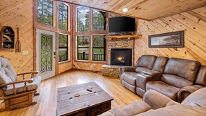 A cozy cabin interior with a stone fireplace, leather sofa, wooden flooring, and large windows offering a view of the surrounding forest. - Honey Hole Cabin is located on the Carson Creek Rd. Minutes to the Lake (Broken Bow)
