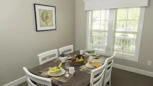 Un coin repas avec une table dressée pour quatre personnes, des chaises blanches, un tableau encadré au mur et une fenêtre avec des stores.