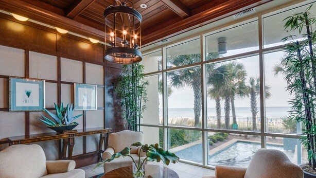 A spacious living area with a large window overlooking a pool and palm trees, featuring a wooden ceiling, comfortable seating, and decorative plants.