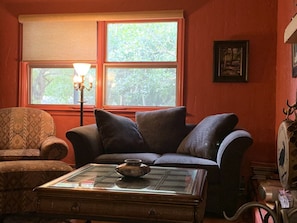 Uma sala de estar acolhedora com uma poltrona estampada, um sofá escuro e uma mesa de centro de vidro com uma tigela decorativa.
