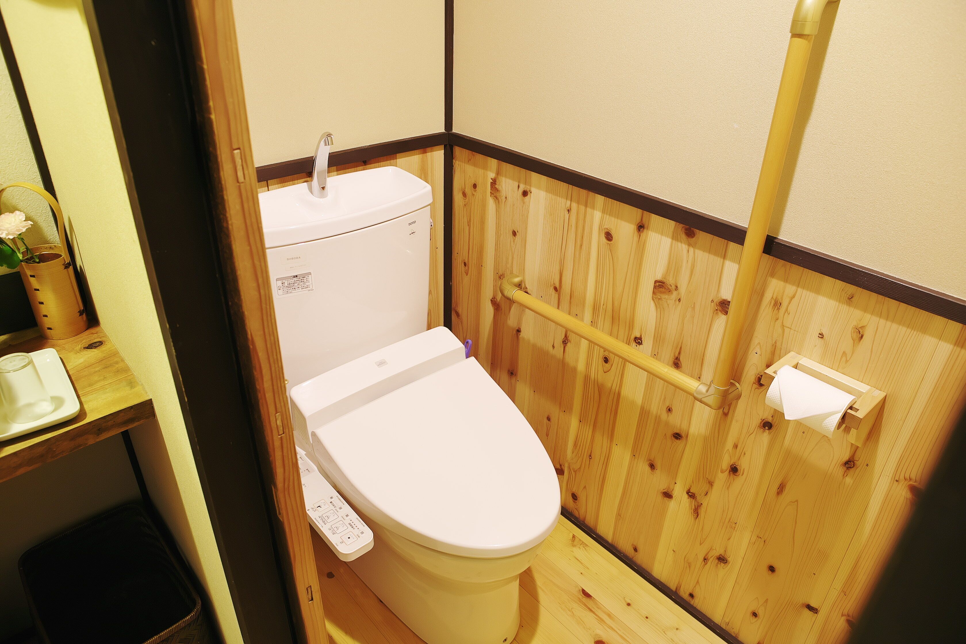 A small, compact bathroom with a wooden wall, a white toilet, a wooden shelf with a glass, and a small trash bin.