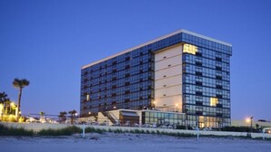 Un hotel moderno con fachada de vidrio, situado cerca de una playa con palmeras.