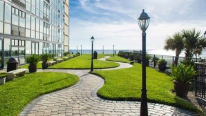Un sendero ajardinado con diseño curvo, bordeado por césped cuidado y plantas en macetas, que conduce a un edificio moderno con amplios ventanales.