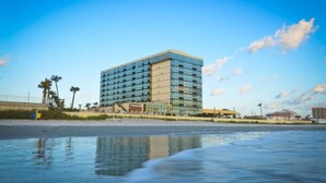 Un hotel moderno con fachada de vidrio, situado cerca de una playa con palmeras y cielo despejado.