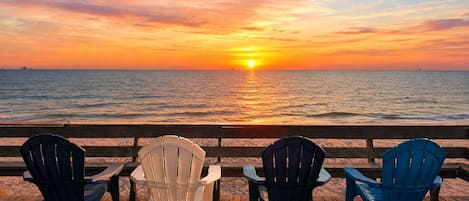 A wooden deck with four Adirondack chairs facing a sunset over the ocean.