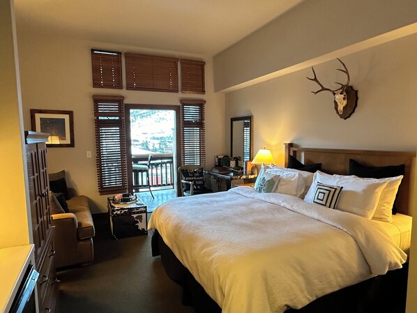 A bedroom with a large bed, a wooden headboard, a mounted deer skull, and a balcony with a view.