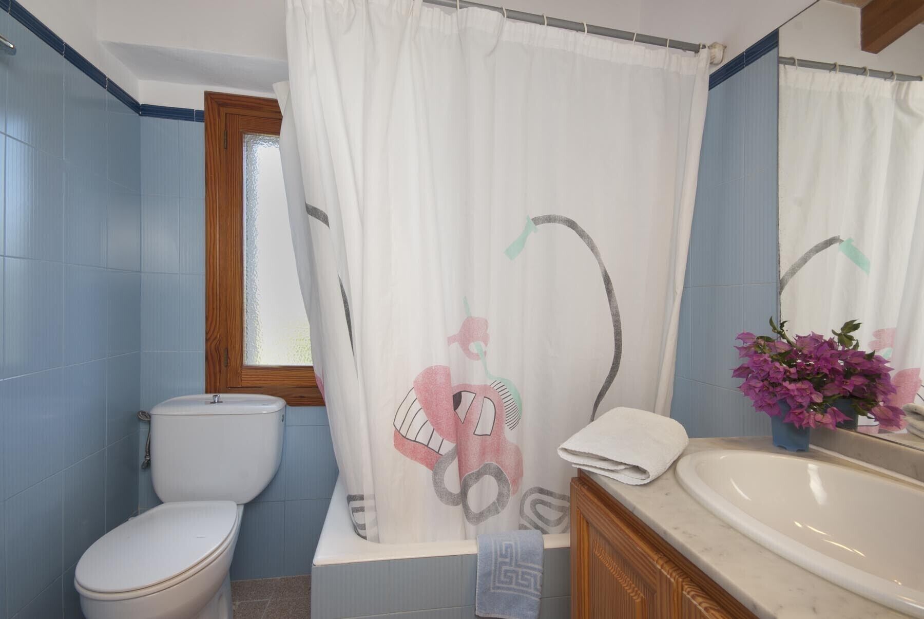 A bathroom with a toilet, a sink, a shower curtain with a design, and a vase with flowers.
