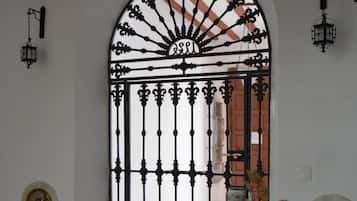 A black wrought iron gate with a fan-like design, a white wall with a circular emblem, and a potted plant with flowers.