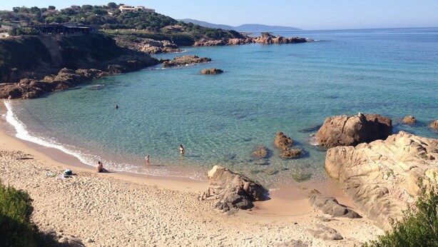 Una playa costera con aguas claras, afloramientos rocosos y una colina distante con edificios.