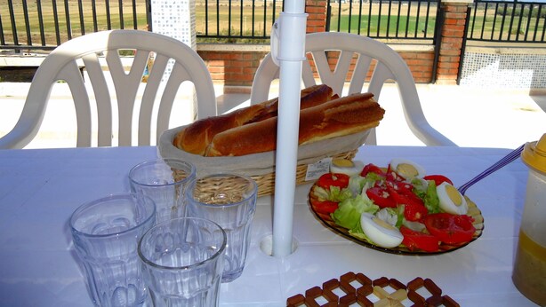 Une table avec un panier de pain, une salade et des verres, installée à l’extérieur, avec vue sur une clôture et de la verdure.