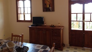 Une pièce avec un meuble en bois, un téléviseur posé dessus, une table avec des chaises et une fenêtre avec des stores.