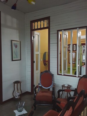 A room with a wooden paneled wall, a door leading to another room, a framed picture on the wall, and two chairs with a small table. - Casa El Mirador - Yliana (Baracoa)